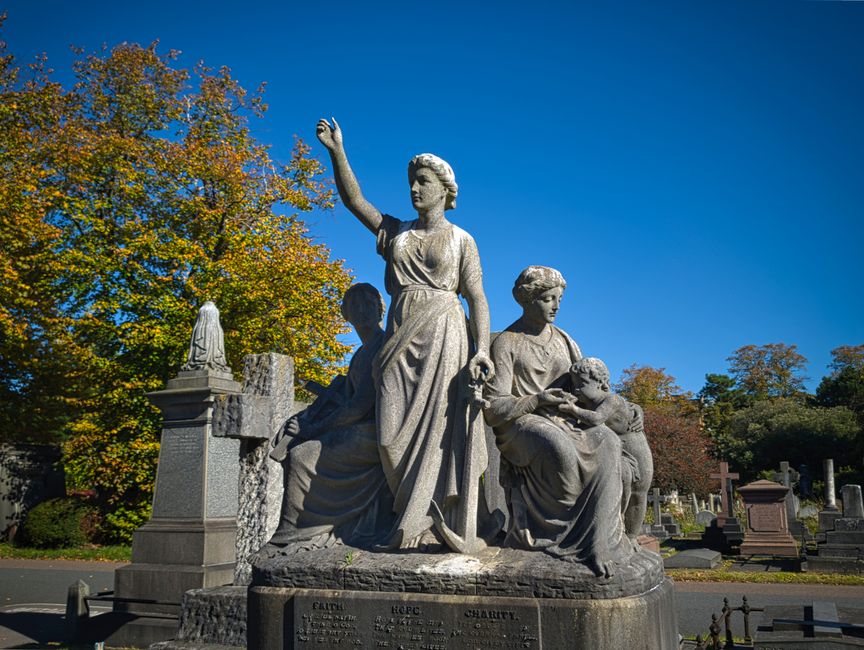 A detailed view of a large stone monument in a cemetery, featuring a central carved female figure with her right arm raised, dressed in flowing robes, standing on a rectangular base. To her left, a seated male figure appears to be resting or contemplating, while to her right, a seated female figure holds a small child in her lap; all three figures are carved from grey stone with intricate detailing. The monument is surrounded by trees with autumn foliage, showing orange, yellow, and green leaves, under a clear blue sky. In the background, additional stone monuments and graves are visible, along with pathways. The scene is lit by natural daylight, emphasizing the textures of the stone carvings and the vibrant colours of the seasonal leaves. This image is associated with house removals or estate clearance, illustrating the careful handling and transportation of memorial monuments, a process sometimes managed by Brompton Man and Van for relocation or preservation services.
