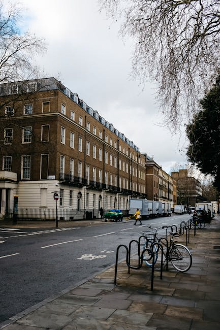 A street scene showing a large, multi-storey brick building with white window frames and small balconies along the upper floors. The building is situated behind a black metal fence and is adjacent to