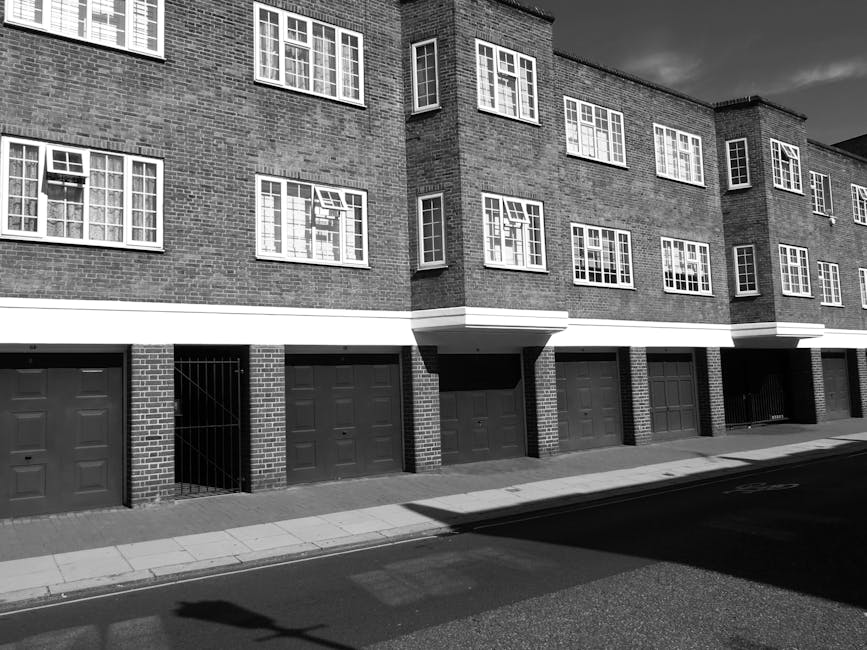 Black and white photograph of a multi-storey residential building on Old Brompton Road with a brick facade and multiple windows, some open, arranged symmetrically on each floor. The ground level features several garage doors in a neat row, with one garage door partially open revealing a gated entryway beside it. The building's entrance is recessed beneath protruding sections of the upper floors. The image captures the building's exterior in daylight, with shadows cast on the pavement and road in the foreground. The scene depicts a typical urban environment suitable for house removals, with the building likely involved in home relocation or furniture transport activities, as facilitated by Brompton Man and Van. The overall setting is clean, orderly, and indicative of residential property management and moving logistics.