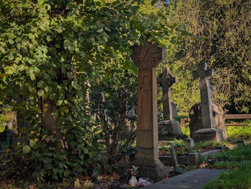 A scene in a graveyard during daytime showing multiple headstones and crosses made of stone and metal, some with intricate carvings, nestled among lush green trees and bushes. The headstones vary in height and design, with one prominent stone cross with detailed carvings in the foreground, partially obscured by tree foliage. The ground is covered with grass and scattered flowers, with some memorial candles placed at the base of a few gravestones. The natural light filters through the leafy canopy, casting dappled shadows across the site. Although the scene is tranquil and well-maintained, the presence of multiple memorials indicate this is a burial ground, part of a site that might be accessed by professional removals or estate services, such as Brompton Man and Van, for cemetery clearances or house removals involving memorial removals.