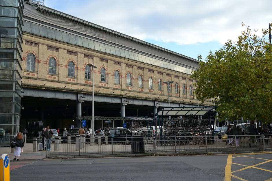 A view of West Brompton station with an overhanging historic brick building featuring arched windows and decorative brickwork, situated above a busy curbside area. The scene includes a bicycle shelter